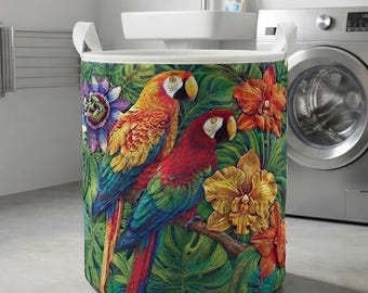 May include: A cylindrical laundry hamper featuring a vibrant tropical print of two macaws, one primarily yellow and orange and the other red and green, surrounded by large green monstera leaves, purple passion flowers, and orange orchids.