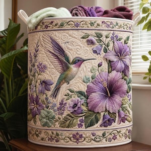 Op de afbeelding: Een cilindrische wasmand van stof met een doorgestikte crèmekleurige achtergrond, voorzien van een geborduurde kolibrie in vlucht, omringd door paarse hibiscus en klokvormige bloemen. De randen hebben een decoratief bloemmotief.