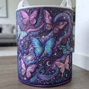 May include: A cylindrical fabric storage basket featuring a quilted design with a vibrant purple and blue celestial background. The exterior is decorated with multiple butterflies in shades of pink, blue, and teal, interspersed with stars, moons, and swirling cosmic patterns. It includes two white fabric handles at the top.