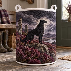 May include: A cylindrical fabric laundry hamper featuring a quilted exterior with a detailed scene of a black dog standing on a rock. The landscape includes purple heather, standing stones, a distant lake, and a stormy sky with a lightning bolt.