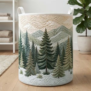 Può includere: Un cesto portaoggetti cilindrico in tessuto con un design trapuntato raffigurante un paesaggio. L'esterno mostra una foresta di alberi sempreverdi in tonalità verde scuro e salvia su un terreno innevato, con montagne e cielo nuvoloso beige.