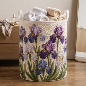 May include: A cylindrical laundry hamper with a cream-colored quilted fabric exterior featuring a vibrant print of purple and white iris flowers with green stems and leaves. The hamper includes two white vertical handles and holds several white towels.