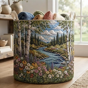 May include: A cylindrical fabric storage basket featuring a quilted landscape print of a winding river, birch trees, pine forests, and colorful wildflowers. The basket has two cream-colored fabric handles and is filled with several folded towels.