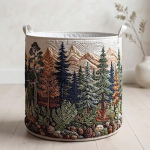 May include: A cylindrical fabric storage basket featuring a quilted texture and a detailed embroidered landscape of a forest with pine trees, ferns, acorns, and mountains in shades of green, brown, blue, and cream, complete with two side handles.
