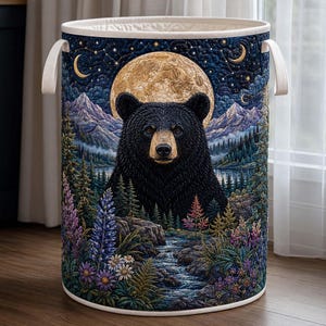 May include: A cylindrical laundry hamper featuring a quilted exterior with a detailed illustration of a black bear centered before a large golden moon, mountain peaks, a flowing stream, pine trees, and colorful wildflowers under a starry night sky.