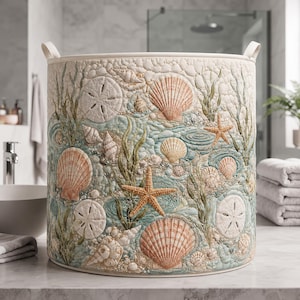 Könnte beinhalten: Ein zylindrischer Wäschekorb aus Stoff mit gesteppter Textur und einem detaillierten Aufdruck von Muscheln, Seesternen und Sanddollars in Beige-, zarten Rosé- und Meergrüntönen. Der Korb verfügt über zwei stabile Stoffgriffe an der Oberseite.