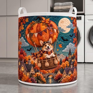 May include: A cylindrical laundry hamper featuring a colorful quilted-style print of a Corgi wearing goggles and a scarf in a pumpkin-shaped hot air balloon. The scene includes a full moon, bats, autumn trees, and a village with stone houses.