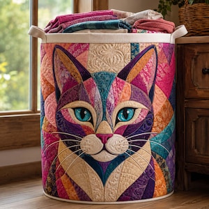 May include: A cylindrical fabric laundry hamper featuring a vibrant, multicolored quilted cat face design. The cat has bright blue eyes, a pink nose, and long white stitched whiskers. The exterior is composed of various geometric fabric patches.
