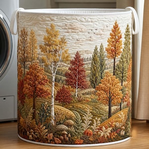 Può includere: Un cesto portabiancheria cilindrico in tessuto con un paesaggio trapuntato raffigurante un bosco autunnale con betulle, pini e alberi a foglia caduca nei toni dell'arancione, giallo, rosso e verde, con due manici bianchi sul bordo.