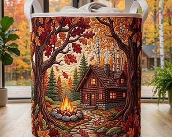 May include: A cylindrical fabric laundry hamper featuring a quilted design of a log cabin in an autumn forest. The scene depicts a stone path leading to a cabin, a campfire, and trees with orange and red leaves. It has two white top handles.
