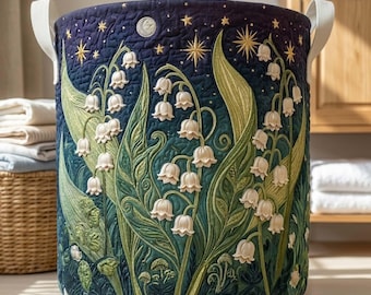 May include: A cylindrical fabric storage basket featuring a quilted design with a dark blue night sky background, white lily of the valley flowers, green leaves, and gold embroidered stars and a crescent moon, complete with two white side handles.
