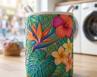May include: A cylindrical laundry hamper featuring a vibrant tropical print with orange bird of paradise flowers, pink hibiscus, and yellow plumeria blossoms set against a background of green monstera leaves and ferns, finished with white rope handles.