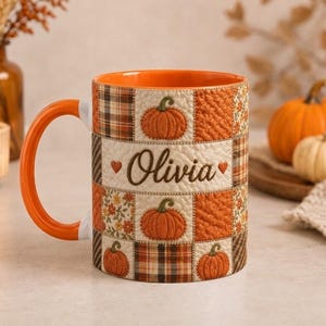 Può includere: Una tazza in ceramica con manico e interno arancioni, caratterizzata da un design patchwork con zucche, motivi a quadri e decorazioni floreali. Il nome Olivia è stampato in corsivo marrone al centro, affiancato da due piccoli cuori.