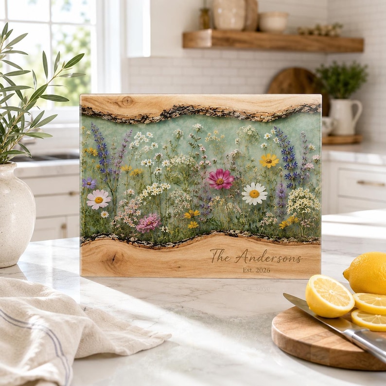May include: A rectangular wooden serving board with natural live edges and a central resin inlay featuring a pressed wildflower meadow design. The board is engraved with the text The Andersons Est. 2026 on the lower right side of the wood.