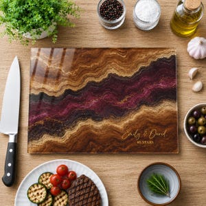 May include: A rectangular glass cutting board featuring a swirling agate-inspired pattern in shades of brown, black, and deep magenta. The board is personalized with the gold text "Emily & David" and "40 YEARS" printed in the lower right corner.