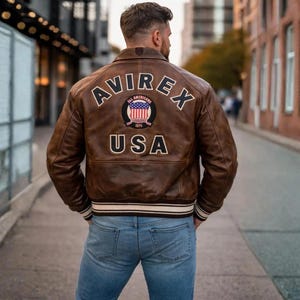 Pode incluir: Uma jaqueta bomber de couro marrom com o texto AVIREX e USA em letras brancas nas costas. Um emblema circular com o design da bandeira americana e o texto ALL AMERICANS ESTABLISHED 1975 está centralizado entre as palavras. Punhos canelados.