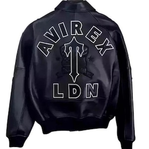 May include: A black leather jacket featuring a large white embroidered logo on the back. The text AVIREX is arched above a central T symbol, with LDN printed below. The jacket has a classic collar, long sleeves, and ribbed cuffs and hem.