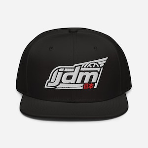 Peut inclure: Une casquette snapback noire avec un motif brodé blanc sur le panneau avant. Le design affiche les lettres jdm dans une police stylisée avec un élément en forme d'aile au-dessus et les caractères japonais rouges 日本 en dessous.