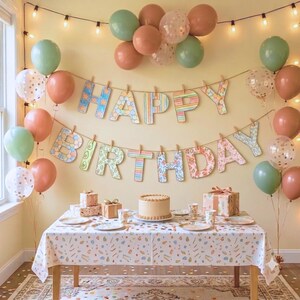May include: A birthday party setup featuring a table with a patterned tablecloth, a frosted cake, and gift boxes. Above the table, a banner spells out HAPPY BIRTHDAY with patterned letters, surrounded by sage green, terracotta, and gold confetti balloons.