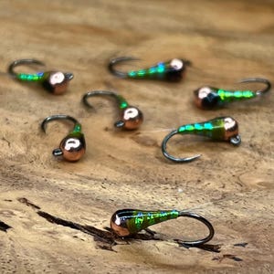 May include: A collection of seven fishing flies, each featuring a metallic copper-colored bead head and a slender body with a vibrant, iridescent green scale pattern. Each fly is equipped with a curved metal hook, designed for fly fishing applications.