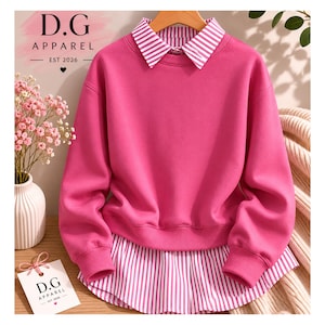 Puede incluir: Una sudadera magenta con cuello y dobladillo a rayas blancas y rosas. La sudadera tiene mangas largas y cuello redondo. El texto "D.G Apparel EST 2026" está impreso en una etiqueta pequeña.