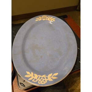 Pode incluir: Uma travessa de servir oval em cerâmica num tom suave de azul pervinca. A travessa apresenta um motivo floral branco com folhas estilizadas e uma flor de rosa central posicionada nas bordas superior e inferior do rebordo.