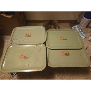 May include: A set of four rectangular light green metal serving trays. Each tray features a small, centred graphic illustration of three figures in orange and yellow clothing. The trays show signs of wear, including surface scuffs and rust spots.