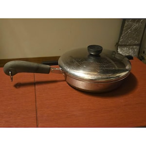 Pode incluir: Uma frigideira redonda de aço inoxidável com acabamento polido e tampa a condizer. A frigideira apresenta um cabo ergonómico longo e cinzento com um pequeno laço de metal na extremidade para pendurar. A tampa tem um botão central preto.