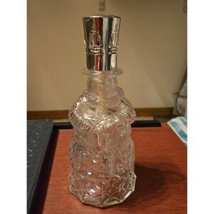 May include: A clear glass fragrance lamp shaped like a snowman wearing a scarf and holding a gift. The lamp features a textured diamond pattern on the base and is topped with a polished silver-colored metal crown with cut-out flame designs.