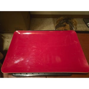 May include: A rectangular serving tray with a vibrant red surface and rounded corners. The tray features a smooth, glossy finish with visible light reflections across the top, and it is positioned horizontally on a dark, flat base.