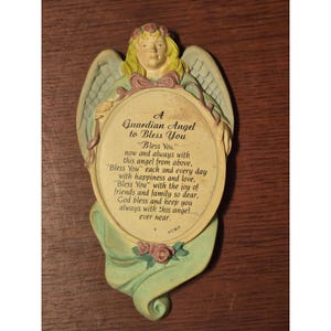 May include: A decorative wall plaque featuring a sculpted angel with blonde hair and light blue wings, framing a central oval panel. The panel contains the text: A Guardian Angel to Bless You, Bless You now and always with this angel from above.
