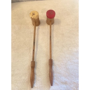 May include: Two long-handled wooden kitchen tools with cylindrical heads. One head is painted cream with visible wear, and the other is painted red. Both feature perforated tops and slender wooden shafts leading to tapered, rounded handles.