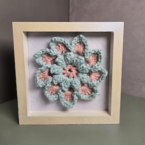 May include: A square light-colored wooden frame containing a crocheted flower motif. The flower features alternating layers of soft sage green and dusty pink yarn, centered on a light gray felt background with subtle glittery flecks throughout.