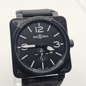 Puede incluir: Un reloj de pulsera de caja cuadrada negra con bisel de fibra de carbono, esfera negra con n&uacute;meros ar&aacute;bigos blancos en las 12, 3 y 9, manecillas blancas, una subesfera en la posici&oacute;n de las 6 y una correa de caucho negra con el grabado BR.