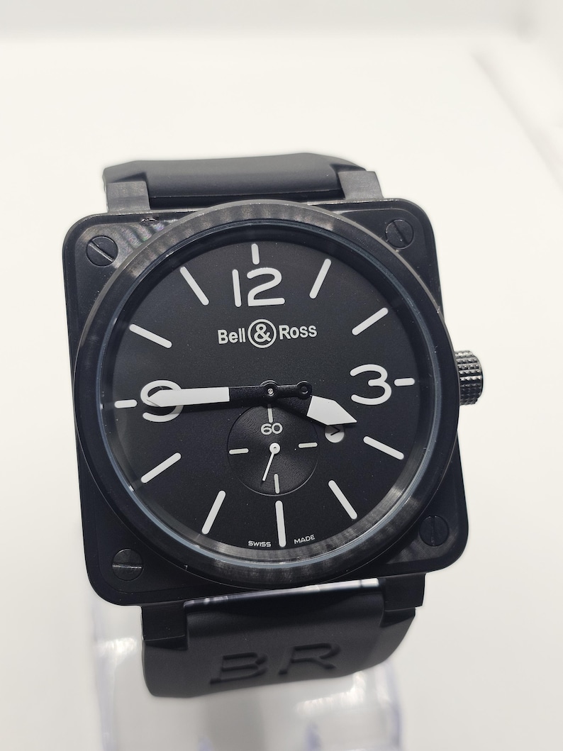 Puede incluir: Un reloj de pulsera de caja cuadrada negra con bisel de fibra de carbono, esfera negra con n&uacute;meros ar&aacute;bigos blancos en las 12, 3 y 9, manecillas blancas, una subesfera en la posici&oacute;n de las 6 y una correa de caucho negra con el grabado BR.