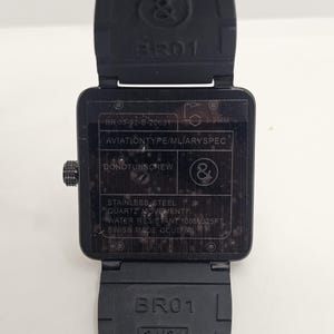 Puede incluir: Un reloj de pulsera cuadrado negro con correa de caucho. La parte posterior de la caja muestra el texto BR 03-92-S-20631, AVIATIONTYPE/MLIARYSPEC, DONOTUNSCREW, STAINLESS STEEL, QUARTZ MOVEMENT, WATER RESISTANT 100M/325FT, SWISS MADE CCUE y el logo.