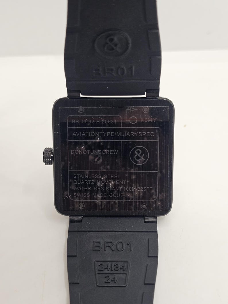 Puede incluir: Un reloj de pulsera cuadrado negro con correa de caucho. La parte posterior de la caja muestra el texto BR 03-92-S-20631, AVIATIONTYPE/MLIARYSPEC, DONOTUNSCREW, STAINLESS STEEL, QUARTZ MOVEMENT, WATER RESISTANT 100M/325FT, SWISS MADE CCUE y el logo.