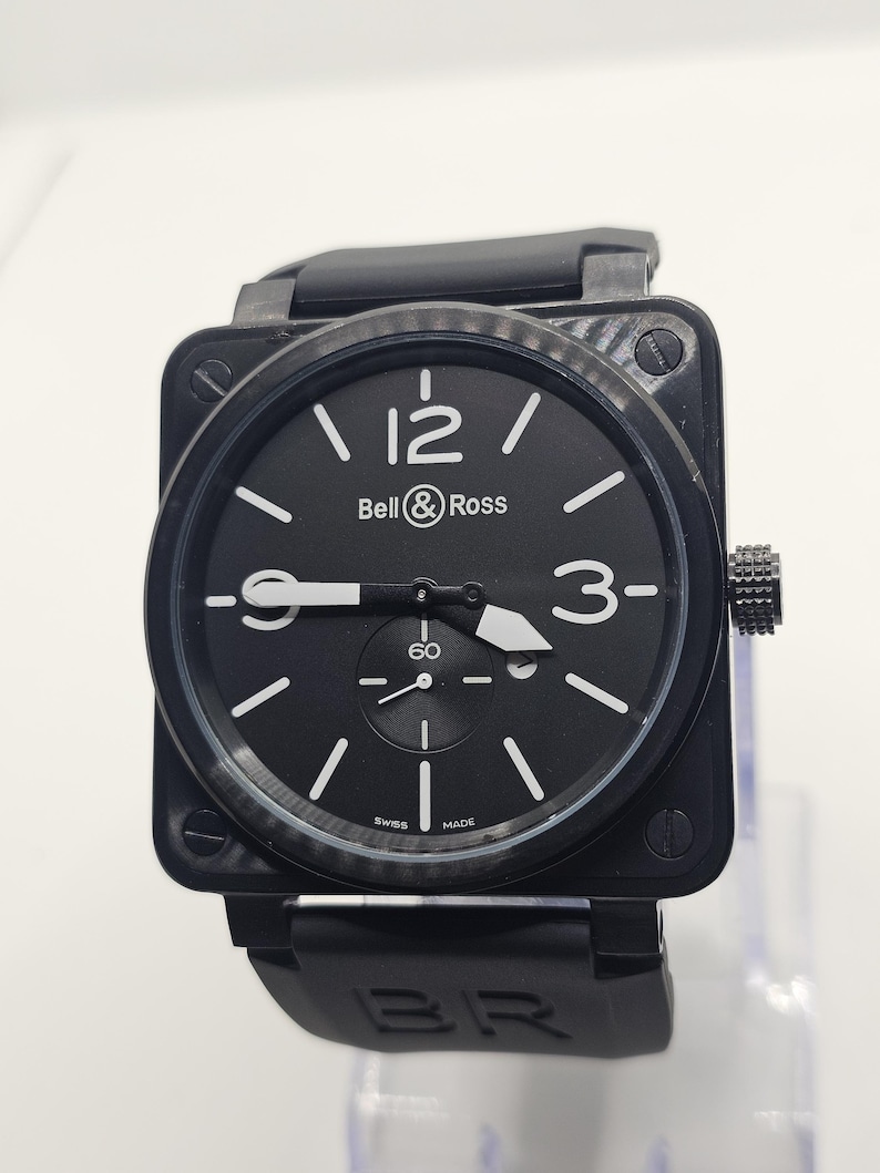 Puede incluir: Un reloj de pulsera con caja cuadrada negra y esfera negra que presenta el logotipo de Bell & Ross, n&uacute;meros ar&aacute;bigos blancos en las 12 y las 3, e &iacute;ndices de bast&oacute;n blancos. El reloj incluye una subesfera en la posici&oacute;n de las 6, manecillas blancas y una correa de caucho negra.