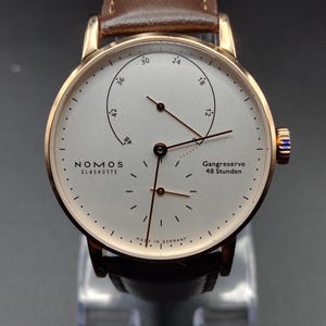 Peut inclure: Une montre-bracelet Nomos Glashütte avec un boîtier couleur or rose, un cadran blanc argenté et un bracelet en cuir marron. Le cadran présente des index noirs, des aiguilles fines, un indicateur de réserve de marche Gangreserve 48 Stunden.