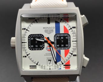Könnte beinhalten: Eine quadratische Monaco Heuer Chronographenuhr mit weißem Zifferblatt, blauen und orangefarbenen vertikalen Streifen, zwei schwarzen Hilfszifferblättern, einem Datumsfenster auf der Sechs-Uhr-Position und einem weißen Lederarmband mit orangefarbenen Nähten.