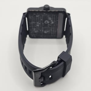 Puede incluir: Un reloj de pulsera negro con esfera cuadrada y correa de caucho negro mate con cierre de hebilla. La hebilla tiene grabadas las letras BR. La caja del reloj presenta una corona texturizada en la parte superior y una esfera oscura y detallada.