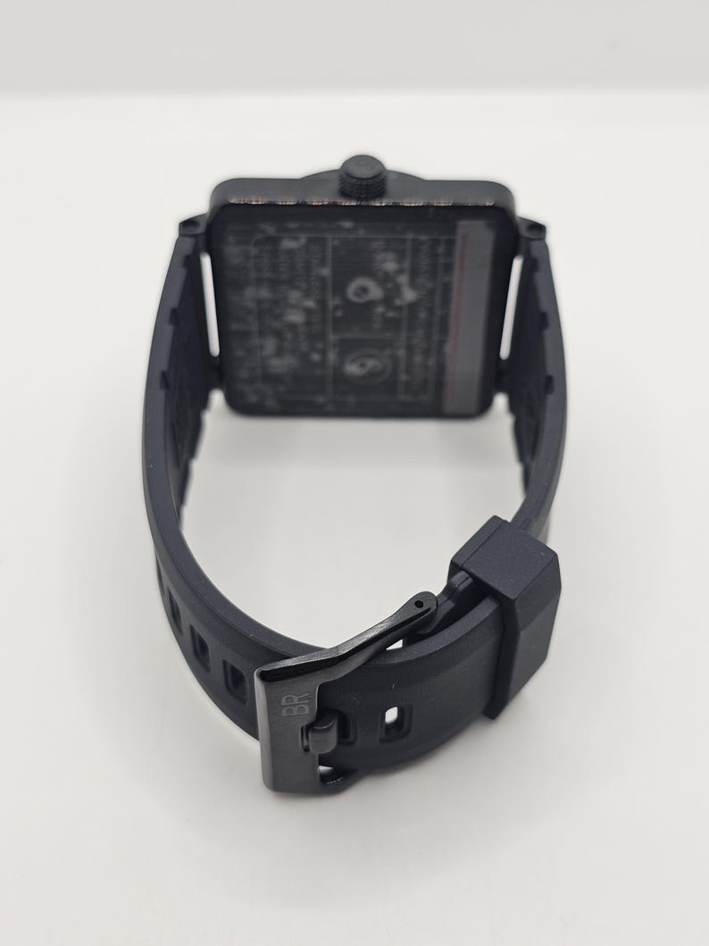 Puede incluir: Un reloj de pulsera negro con esfera cuadrada y correa de caucho negro mate con cierre de hebilla. La hebilla tiene grabadas las letras BR. La caja del reloj presenta una corona texturizada en la parte superior y una esfera oscura y detallada.