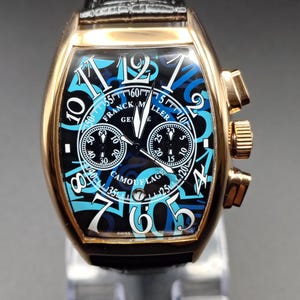 May include: A Franck Muller wristwatch with a rose gold tonneau-shaped case and a black leather strap. The dial features a blue and black camouflage pattern, large white Arabic numerals, two sub-dials, a date window, and the text Franck Muller Geneve.