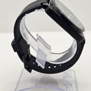 Puede incluir: Un reloj de pulsera negro con caja redonda y correa de silicona negra lisa. El reloj tiene una esfera oscura con manecillas y marcadores visibles, asegurado con un cierre de hebilla est&aacute;ndar. El dise&ntilde;o es monocrom&aacute;tico, mostrando un acabado negro mate elegante en todo el reloj, incluyendo la caja, la correa y los componentes de la hebilla.