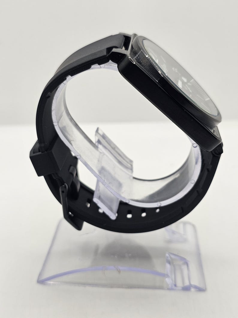 Puede incluir: Un reloj de pulsera negro con caja redonda y correa de silicona negra lisa. El reloj tiene una esfera oscura con manecillas y marcadores visibles, asegurado con un cierre de hebilla est&aacute;ndar. El dise&ntilde;o es monocrom&aacute;tico, mostrando un acabado negro mate elegante en todo el reloj, incluyendo la caja, la correa y los componentes de la hebilla.