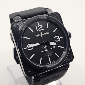 Puede incluir: Un reloj de pulsera de caja cuadrada negra con una esfera redonda negra que presenta n&uacute;meros ar&aacute;bigos blancos en las 12, 3 y 9. La esfera muestra el texto Bell & Ross y Swiss Made, con una peque&ntilde;a subesfera en la posici&oacute;n de las 6 y correa de caucho negro.