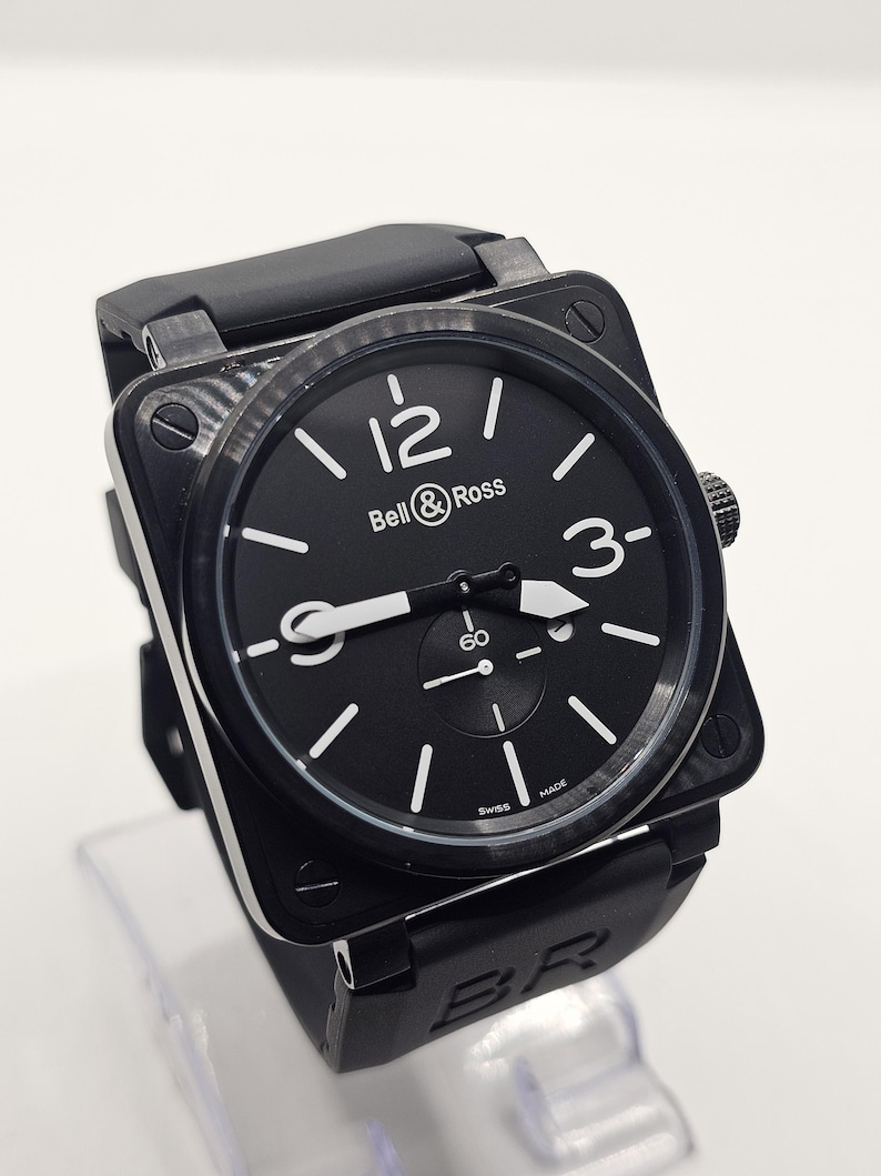 Puede incluir: Un reloj de pulsera de caja cuadrada negra con una esfera redonda negra que presenta n&uacute;meros ar&aacute;bigos blancos en las 12, 3 y 9. La esfera muestra el texto Bell & Ross y Swiss Made, con una peque&ntilde;a subesfera en la posici&oacute;n de las 6 y correa de caucho negro.
