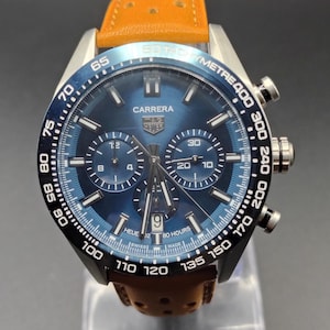 May include: A TAG Heuer Carrera chronograph watch with a blue sunray dial, three sub-dials, and a date window at the six o'clock position. The watch features a black tachymeter bezel, silver-tone hands and indices, and a perforated orange leather strap.