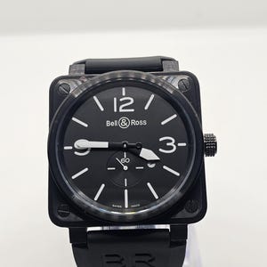 Puede incluir: Un reloj Bell & Ross de caja cuadrada con esfera negra, n&uacute;meros blancos en las 12 y las 3, y una subesfera en la posici&oacute;n de las 6. El reloj presenta un bisel de fibra de carbono texturizado, manecillas luminosas blancas y una correa de caucho negra con el grabado BR.