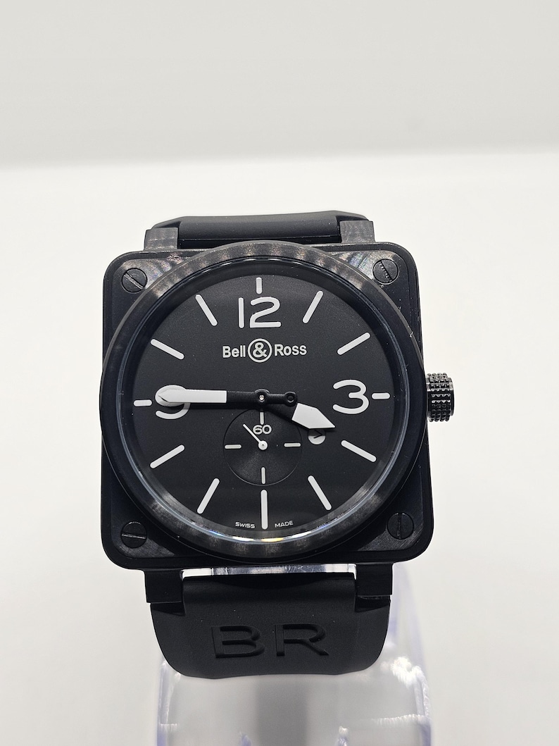 Puede incluir: Un reloj Bell & Ross de caja cuadrada con esfera negra, n&uacute;meros blancos en las 12 y las 3, y una subesfera en la posici&oacute;n de las 6. El reloj presenta un bisel de fibra de carbono texturizado, manecillas luminosas blancas y una correa de caucho negra con el grabado BR.